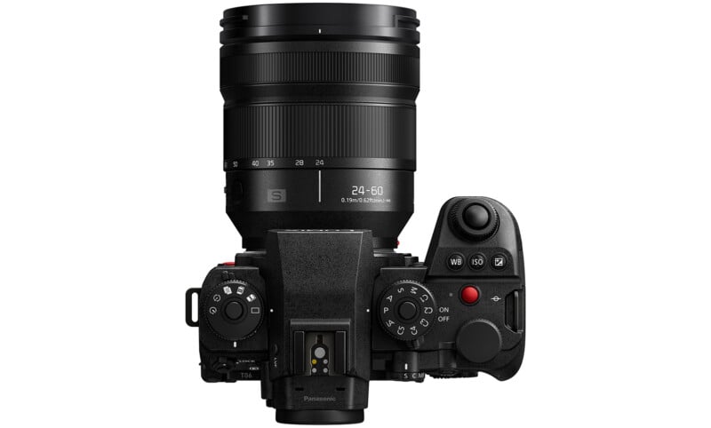 Top view of a black digital mirrorless camera with a zoom lens attached, showing various control dials, buttons, and the on/off switch. The lens markings indicate a 24-60mm focal length.