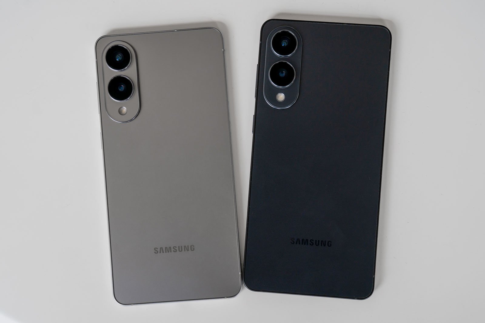 Two Samsung smartphones are placed side by side on a white surface. Both have dual rear cameras aligned vertically in the top left corner. One phone is gray and the other is black.