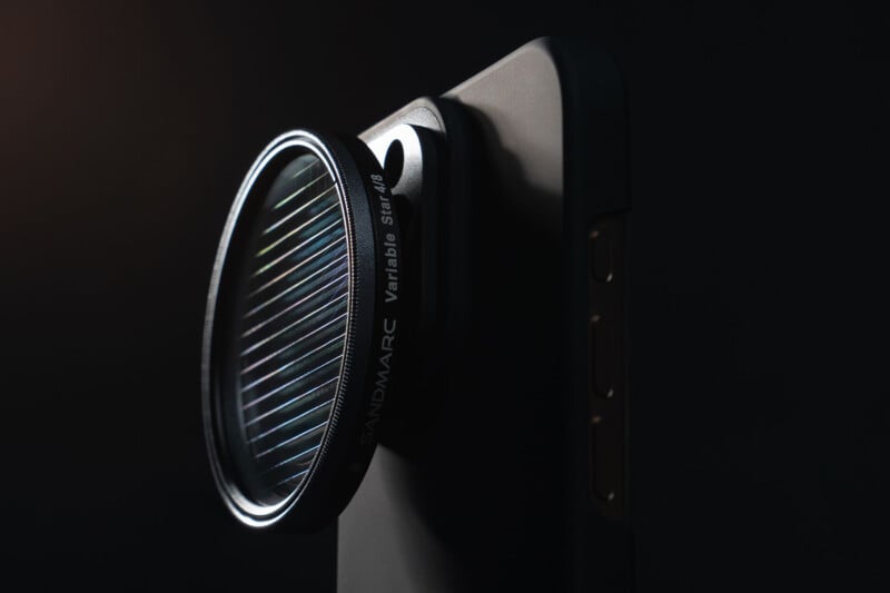 A close-up of a smartphone with a Sandmarc variable star filter attached to its camera, set against a dark background with dramatic lighting highlighting the lens and filter details.