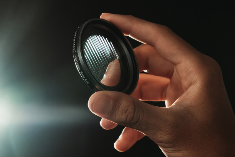 A hand holds a camera lens filter with engraved lines, illuminated by a bright light source from the left against a dark background.