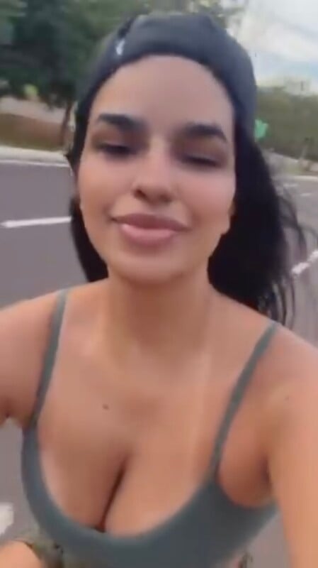A woman with long dark hair wearing a dark tank top and a backward cap smiles while outdoors on a road, with trees and pavement visible in the background.