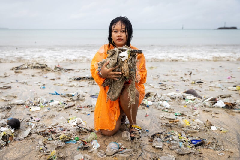 A woman in an orange raincoat kneels on a littered beach, holding a large bundle of trash, with more plastic and debris scattered around her and the shoreline visible in the background.