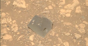 A smooth, gray, glassy rock with angular edges sits among rough, beige, and dusty rocks and pebbles on a sandy surface.