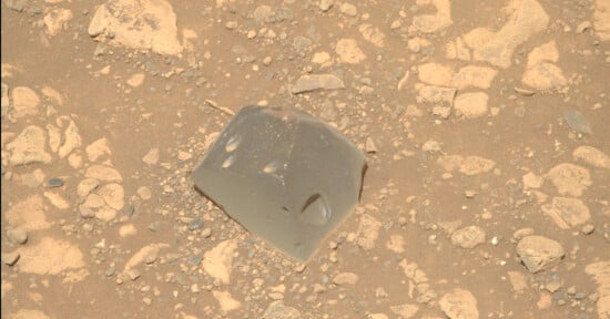 A smooth, gray, glassy rock with angular edges sits among rough, beige, and dusty rocks and pebbles on a sandy surface.