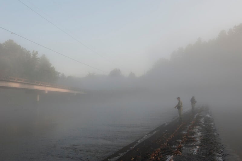 Two people fish in a river on a foggy morning. A bridge is visible on the left, and trees are faintly seen through the mist in the background. The scene appears calm and quiet with low visibility.