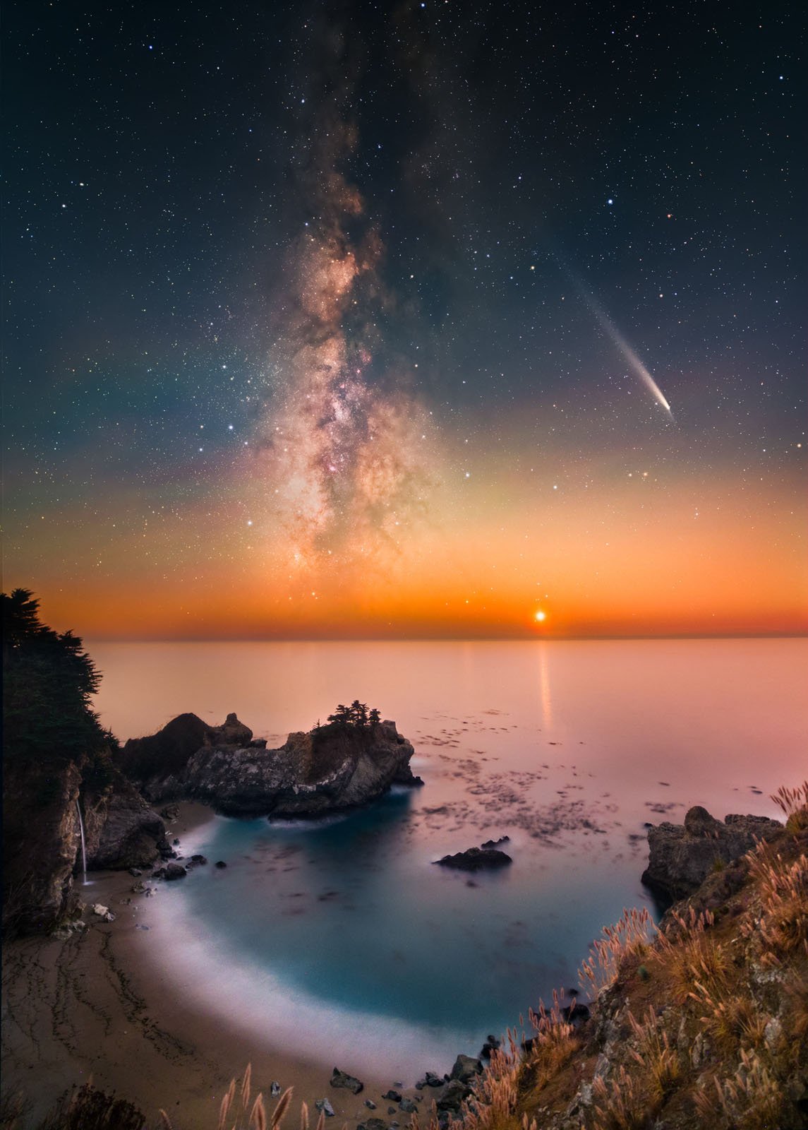 A rocky coastline at twilight under a star-filled sky, with the Milky Way and a bright comet visible above the calm sea. The horizon glows orange as the sun sets, and gentle waves wash onto the sandy shore below.