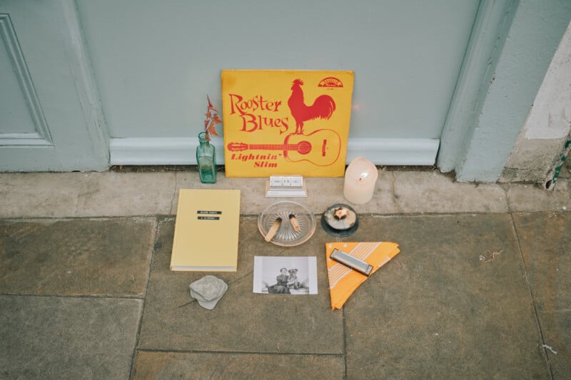 A collection of items on the ground in front of a blue door, including a Rooster Blues vinyl record, a candle, a book, a small glass bottle, a photo, a rock, a plate of shells, and a folded orange cloth.