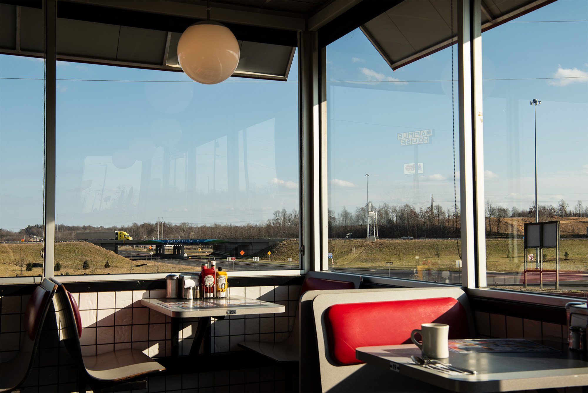 A sunlit diner booth with red seats and a table set with condiments and mugs, next to large windows overlooking a highway, grassy fields, and a clear blue sky.