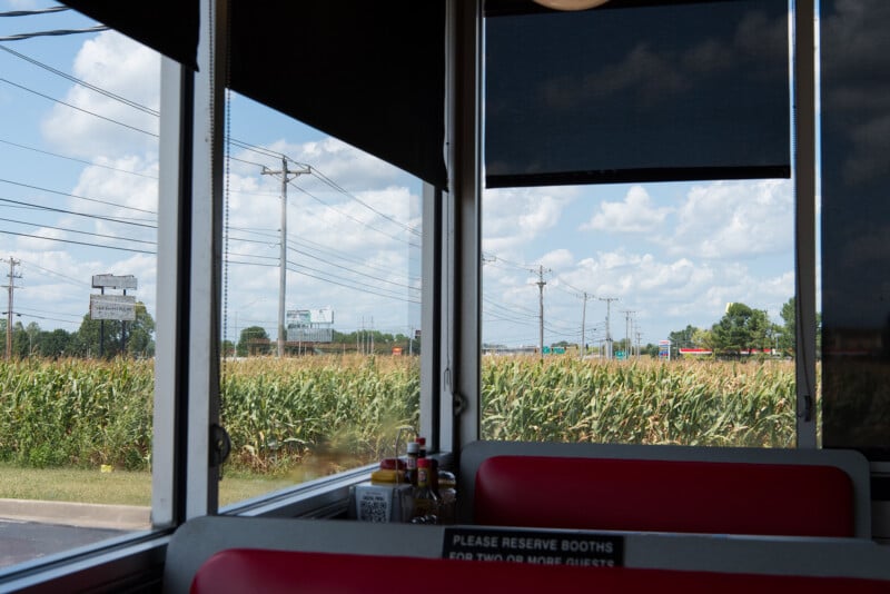 View from inside a diner with red booths, looking out large windows at a field of tall corn and power lines under a bright blue sky with scattered clouds. Condiments are visible on the table.