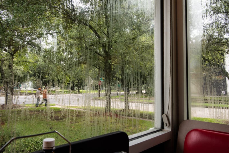 A view through a large, foggy window streaked with condensation, showing two people walking outside on a tree-lined street. Inside, part of a red booth and condiment holders are visible.