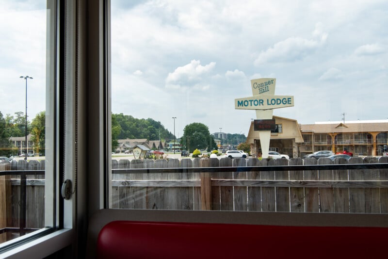 View through a window of a diner with a red seat in the foreground, showing a parking lot, wooden fence, cars, and a retro sign reading "Conner Hill Motor Lodge" under a partly cloudy sky.