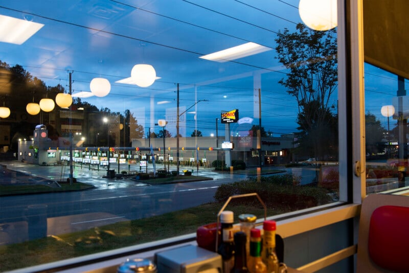 View from inside a diner looking out a window at a Sonic Drive-In across the road at dusk, with reflections of hanging lights and condiment bottles visible on the window.