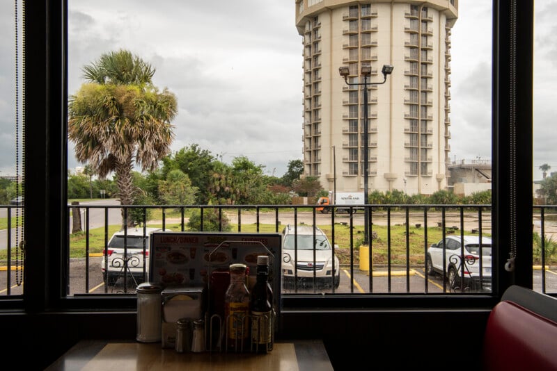 View from inside a diner, looking out a window at parked cars, palm trees, and a tall, round, beige building; diner condiments and a menu are on the table in the foreground.