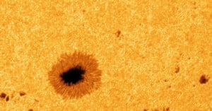 A close-up image of the sun’s surface shows a dark sunspot surrounded by a textured, granular orange-yellow area, highlighting solar activity and surface details.