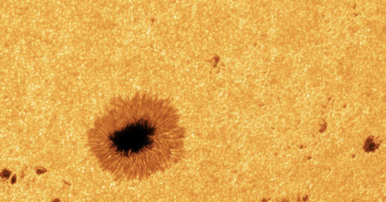 A close-up image of the sun’s surface shows a dark sunspot surrounded by a textured, granular orange-yellow area, highlighting solar activity and surface details.