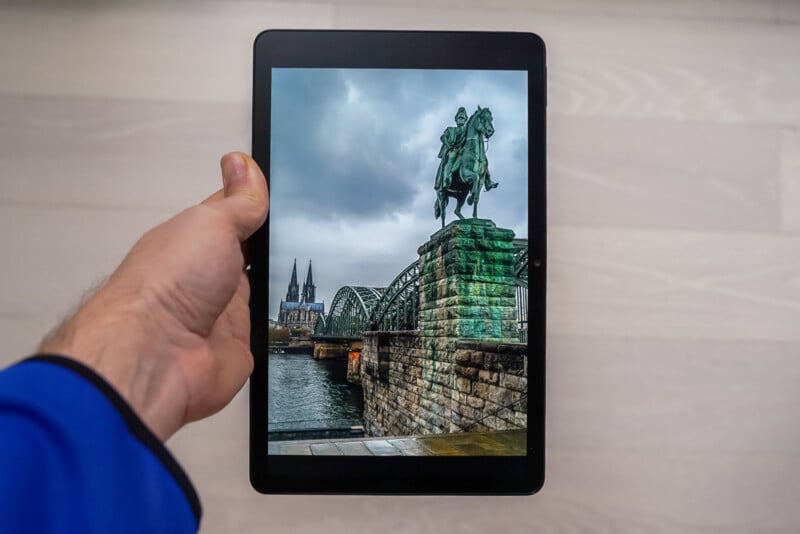 A hand holds a tablet displaying a photo of a riverside statue of a rider on horseback, a bridge, and cathedral towers under a cloudy sky, blending into the real background.