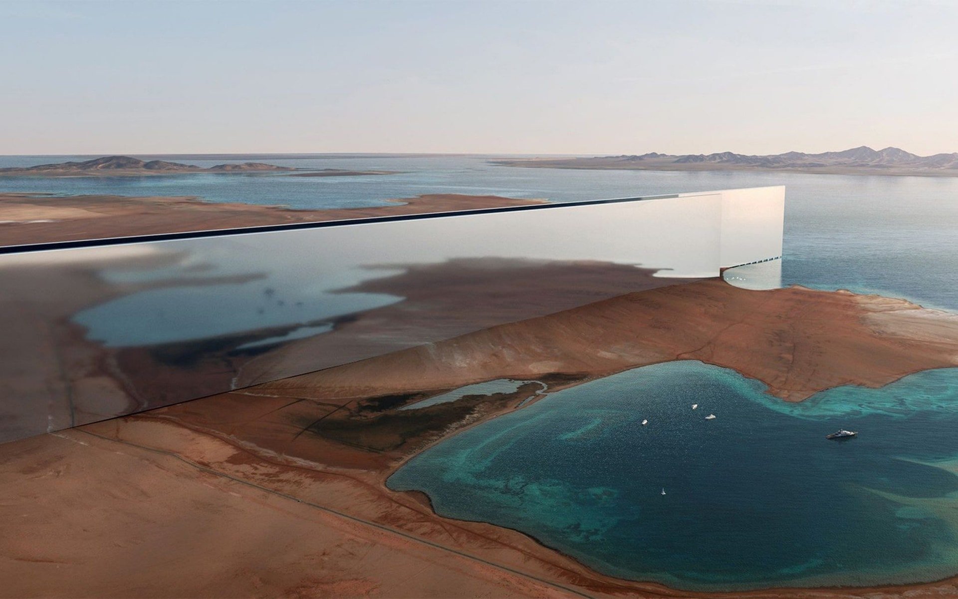 A mirrored, rectangular building stretches across a desert landscape next to the sea, reflecting the sky and surroundings, with mountains and shallow turquoise waters visible in the background.
