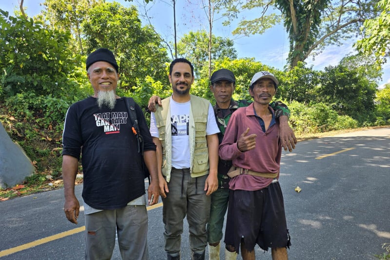 Four men stand together smiling on a paved road surrounded by lush greenery and trees. The weather appears sunny and bright, and the group looks relaxed and happy.