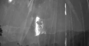 A black and white image shows a bright eruption or explosion with sparks and smoke against a dark sky, partially obscured by streaks of light, possibly from rain or camera artifacts. Trees and mountains are silhouetted below.