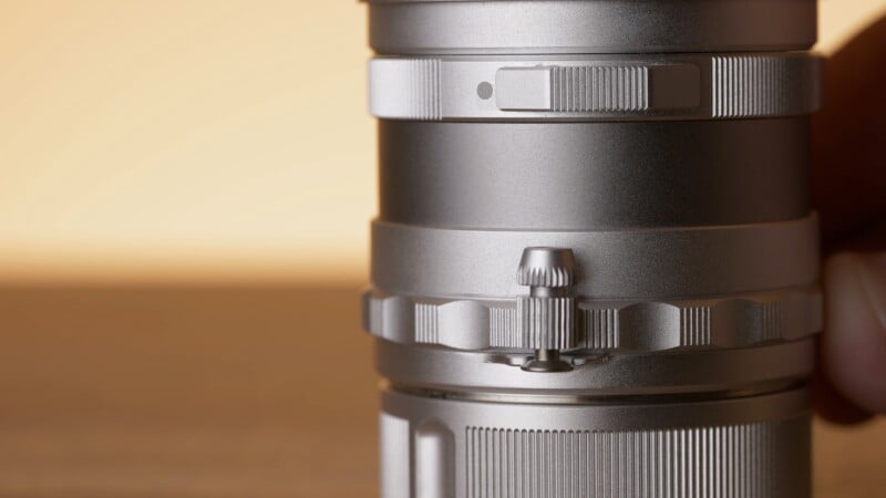 Close-up view of a silver camera lens with textured adjustment rings and knobs, set against a softly blurred, warm-toned background.