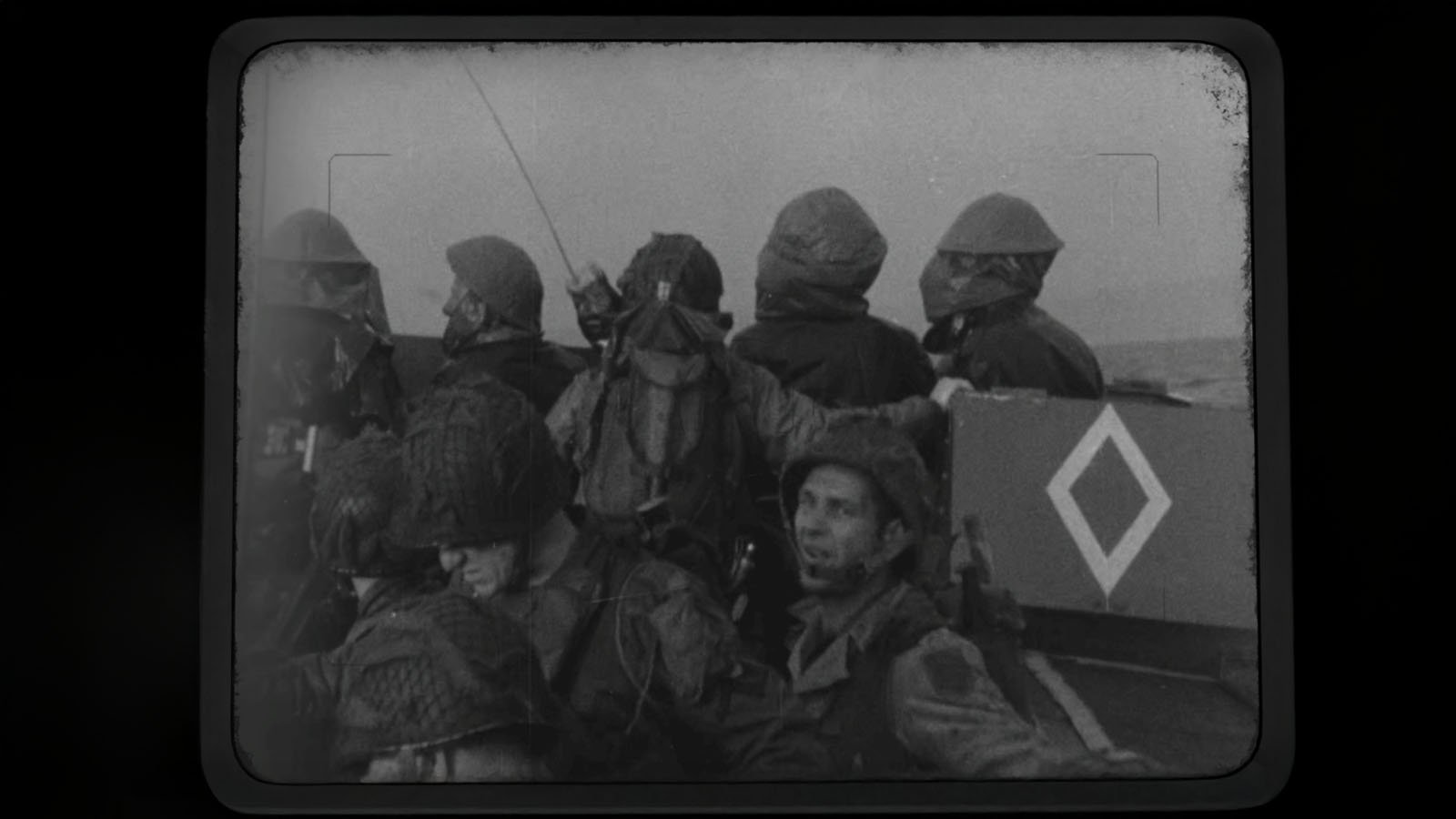 Black and white photo of soldiers wearing helmets and gear in a boat; one soldier holds a radio. The boat has a diamond-shaped symbol on its side. The image appears to be from an old film or footage.