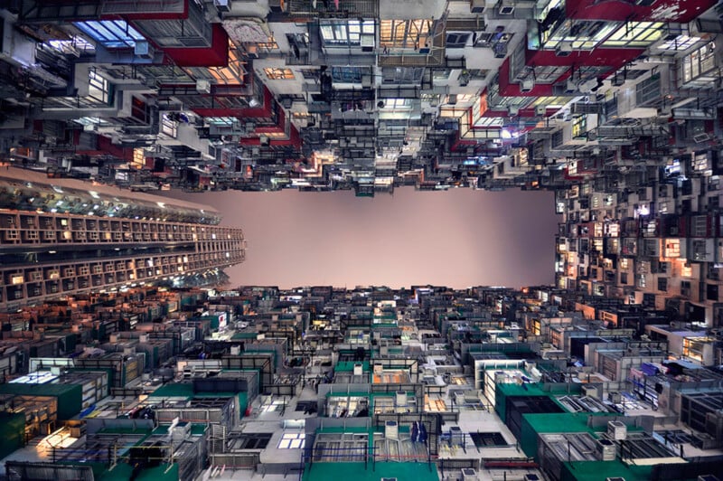 Looking up from a narrow courtyard, densely packed apartment buildings with colorful balconies and windows rise on all sides, forming a tunnel-like effect with a bright, cloudy sky above.