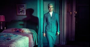 A woman with short blonde hair in a gray suit stands in a dimly lit bedroom, casting a shadow on the wall. The room has a green bed with a floral cover, a bedside lamp, and dark-colored walls and doors.