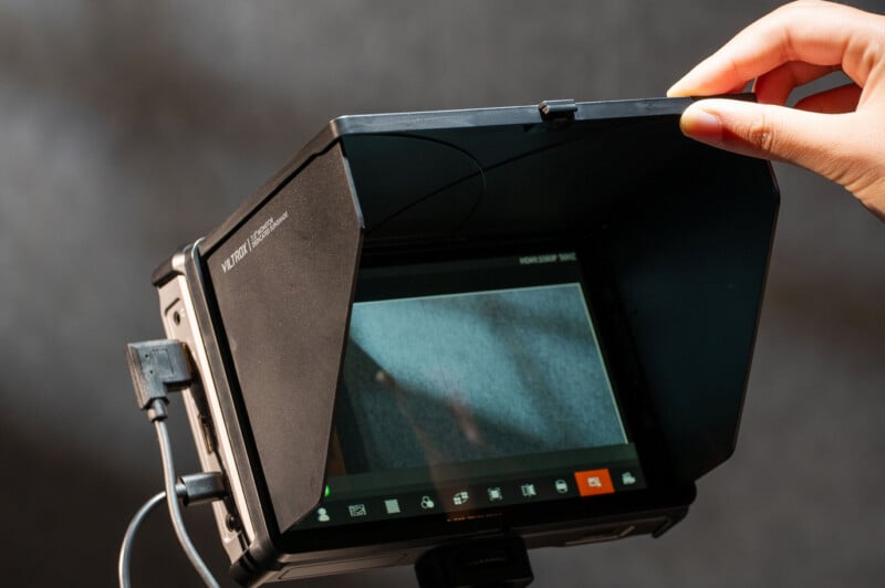A hand lifts the sun hood of a camera monitor, revealing the screen displaying a close-up image. Several cables are connected to the monitor, and camera control icons are visible at the bottom of the screen.