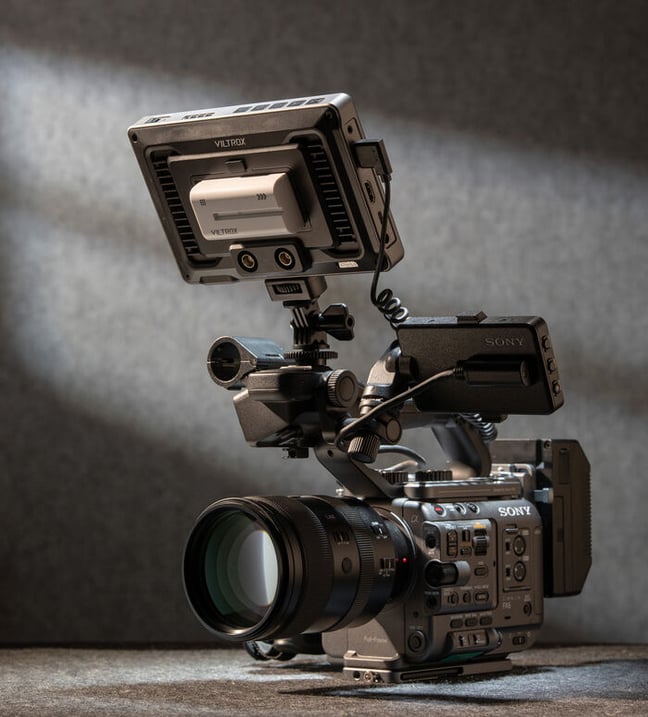 A professional Sony video camera with a large lens and an attached Viltrox monitor sits on a gray surface, lit from the side against a plain gray background.