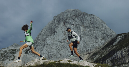 Two people wearing athletic gear and backpacks run or hike on rocky terrain, with a large, steep mountain and cloudy sky in the background.