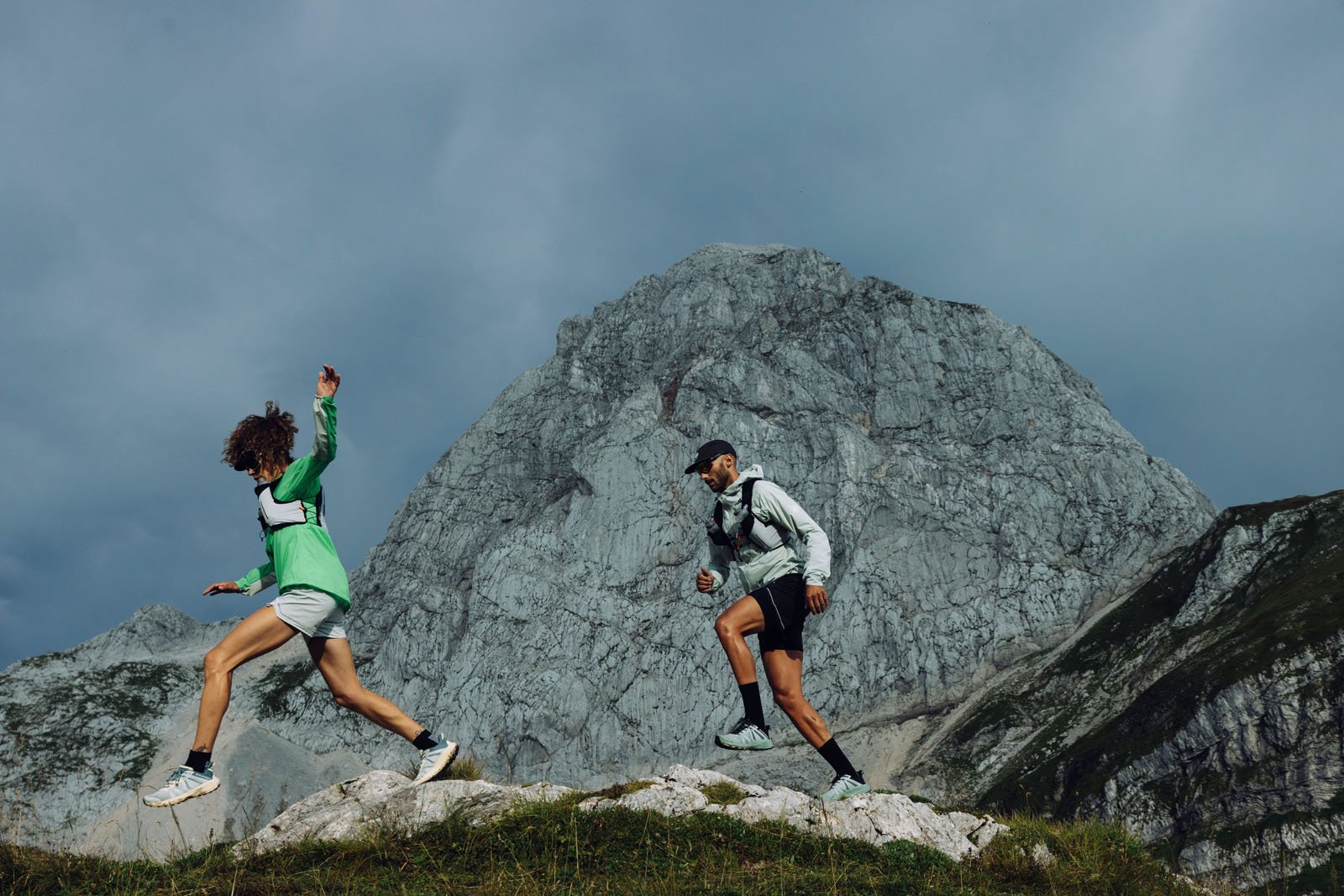 Two people in athletic clothing hike energetically on a rocky trail with a large, rugged mountain in the background under a cloudy sky.