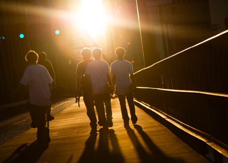 Four people walk on a bridge at sunset, casting long shadows on the ground. The sunlight shines brightly in the background, creating a warm and dramatic atmosphere. One person appears to be holding a skateboard.