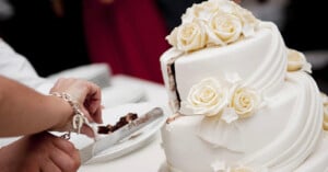 A person cuts a slice of chocolate cake from a white, tiered wedding cake decorated with white icing and white fondant roses, placing the slice on a plate.