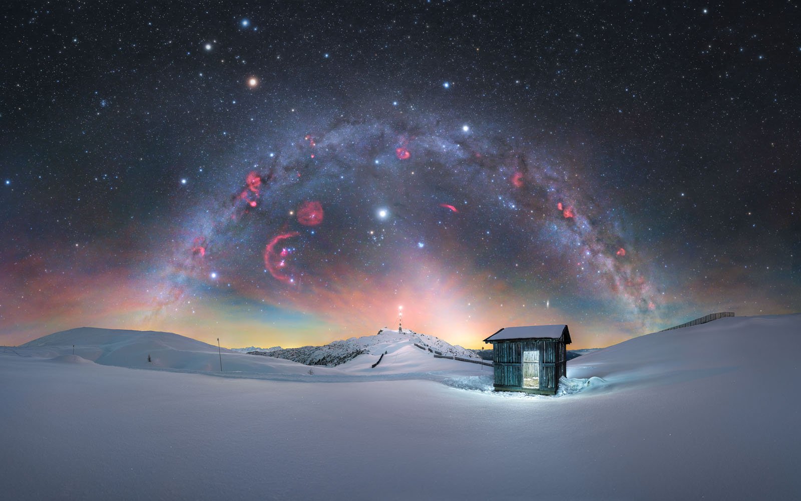 A small shed stands on snowy ground under a starry night sky, with a glowing arc of colorful nebulae and stars stretching above distant hills and a lit structure atop a hill.