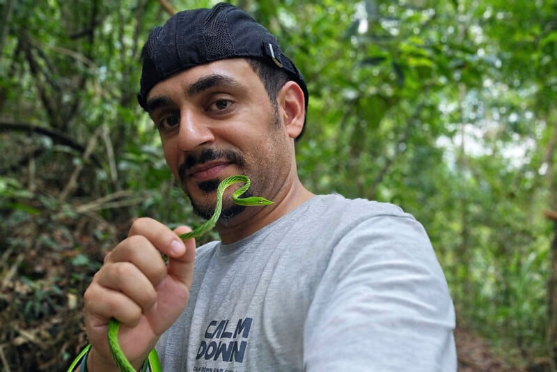 A man in a light gray shirt and black cap holds a small green snake close to his face while standing in a lush, green forest. He looks at the camera with a slight smile, surrounded by dense foliage.