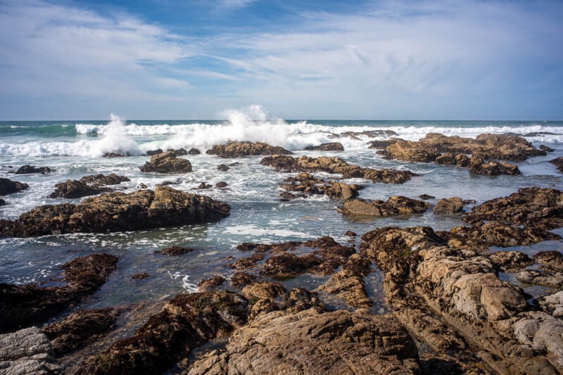 Rocky shoreline with ocean waves crashing against the rocks under a partly cloudy blue sky. Seaweed and wet stones are visible in the foreground.