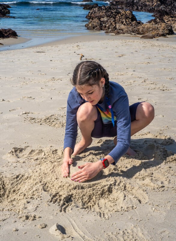 A girl with braided hair kneels on a sandy beach, wearing a blue shirt and shorts, shaping sand with her hands near the shoreline. Rocky outcrops and ocean waves are visible in the background.