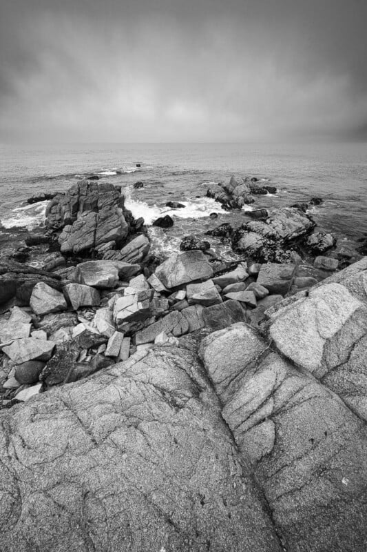 Black and white photo of rocky cliffs leading to an ocean, with waves splashing against jagged rocks under a cloudy sky. The scene appears calm and desolate.