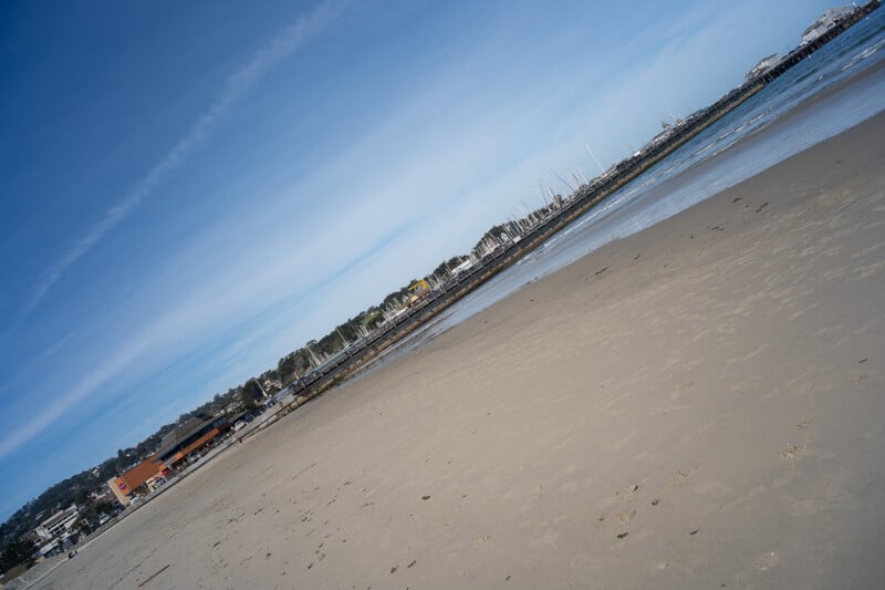 A sandy beach with buildings, a pier, and boats in the background, photographed at a tilted angle under a clear blue sky.