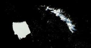 A large rectangular iceberg floats near a rugged, snow-covered island in a dark expanse of ocean, viewed from above.