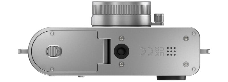 Top view of a silver digital camera showing the lens, battery compartment door with a lock, two screws, and various certification symbols on a smooth metallic surface.