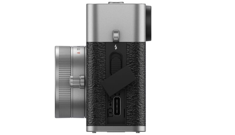 Side view of a digital camera with a textured black and silver body, showing a lens on the left and two exposed ports under a small black rubber cover on the right side.