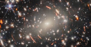 A dense galaxy cluster with bright stars, distant galaxies, and orange arcs of light caused by gravitational lensing against a dark space background.