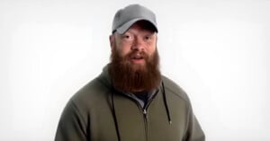 A man with a thick brown beard and mustache wears a gray baseball cap and an olive green hoodie. He stands in front of a plain white background, looking toward the camera.