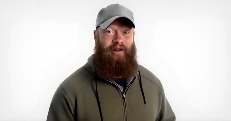 A man with a thick brown beard and mustache wears a gray baseball cap and an olive green hoodie. He stands in front of a plain white background, looking toward the camera.
