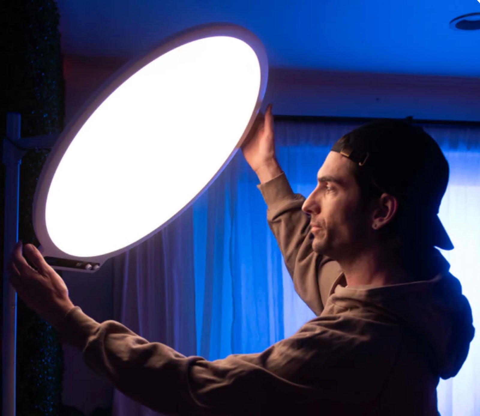 A person wearing a hoodie and a backward cap adjusts a large, circular studio light that glows brightly in a dimly lit room with blue ambient lighting and curtains in the background.
