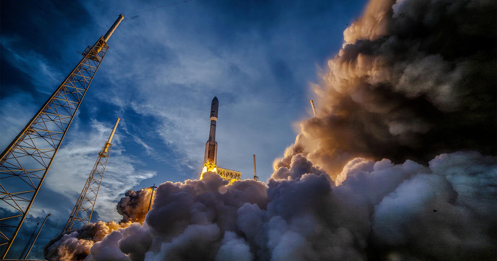 A rocket launches into the sky, surrounded by billowing clouds of smoke and steam, with a dramatic blue and cloudy evening sky in the background and launch towers on either side.