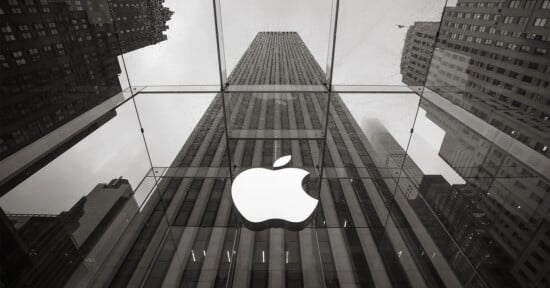 A monochrome photo of the Apple Store entrance featuring a large illuminated Apple logo on glass, with tall city buildings and a cloudy sky reflected in the background.