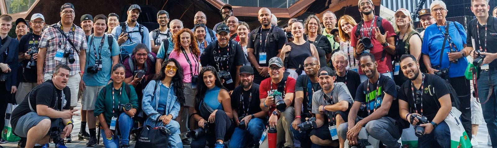 A large, diverse group of people pose together outdoors, many smiling and holding cameras or camera gear, suggesting a photography event or workshop. Everyone is gathered closely in front of a building.