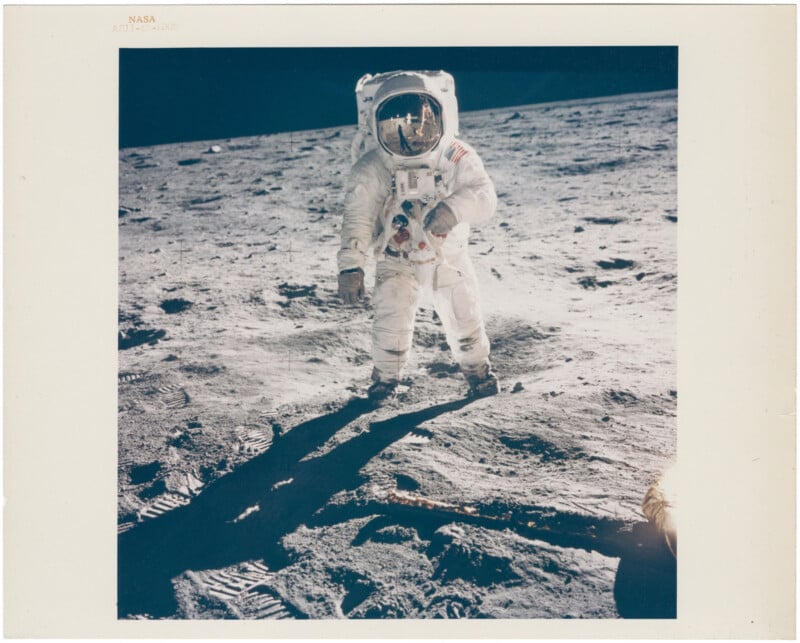 An astronaut in a bulky white spacesuit stands on the rocky, gray surface of the Moon, with shadows and footprints visible in the foreground under a black sky.